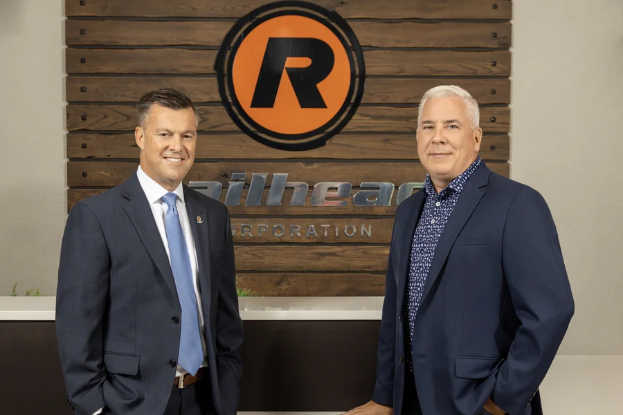 Two men in business attire standing in front of a company sign.