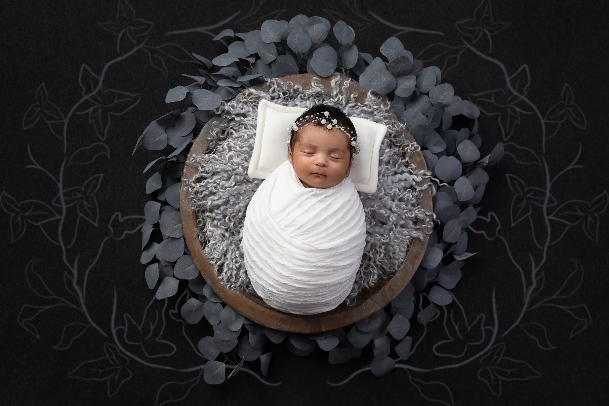 Newborn in white wrap in wooden bowl with gray eucalyptus, dark background — IDTPhoto Chicagoland