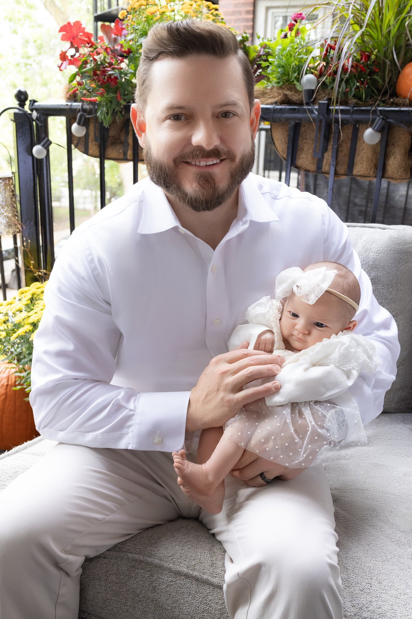Father holding newborn girl in lace dress on outdoor couch surrounded by fall flowers — IDTPhoto Chicagoland lifestyle newborn